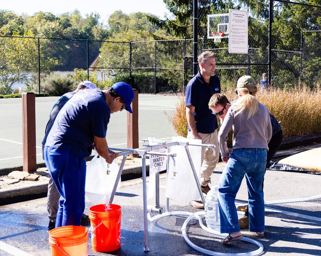 People filling water containers from a temporary water station set up near a tennis court on a sunny day.