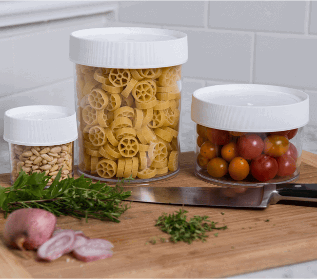 Three clear containers with white lids holding pine nuts, pasta, and cherry tomatoes on a wooden cutting board.