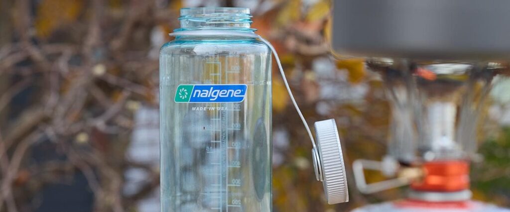 Clear Nalgene water bottle on a table with dry ramen noodles and a camping stove nearby, ready for boiling water outdoors.
